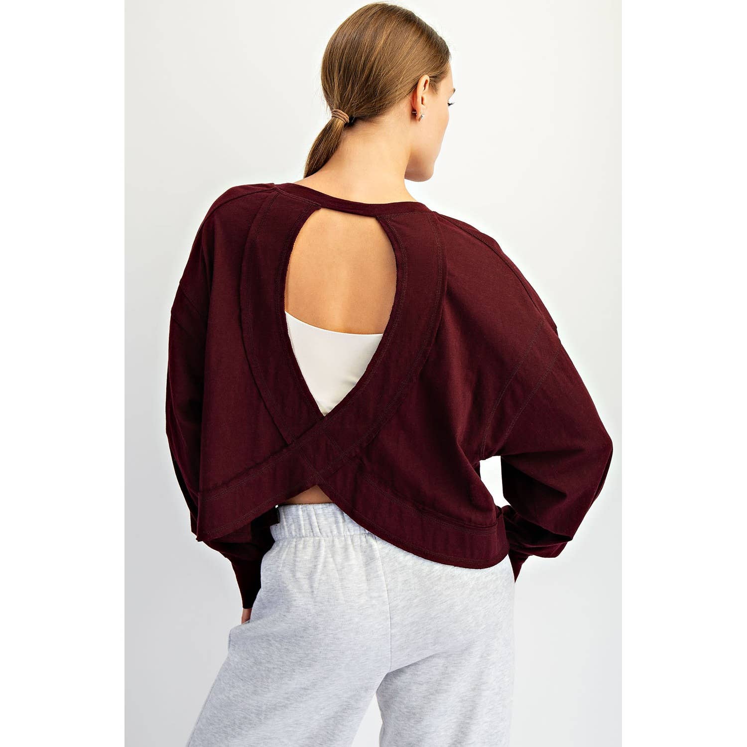 Woman wearing a burgundy open-back top and white pants on a light gray background