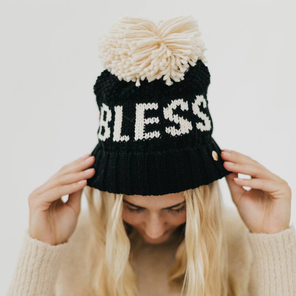 Black knit beanie with white text and pom-pom held by a person against a light background
