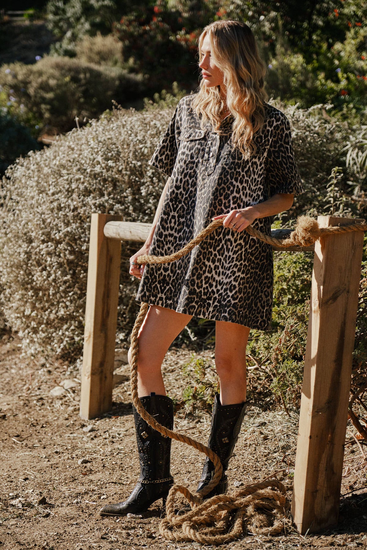 Woman in leopard print dress and boots standing outdoors near wooden posts with rope.