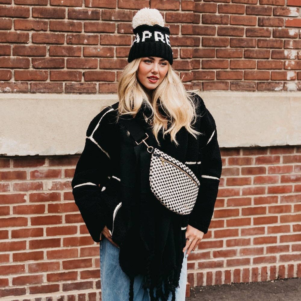 Woman wearing a black jacket, jeans, and a beanie with a checkered bag against a brick wall.