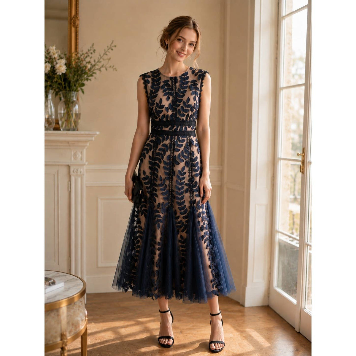 A woman models a sleeveless navy blue lace and tulle midi dress in a bright, elegant room with a fireplace and large windows.