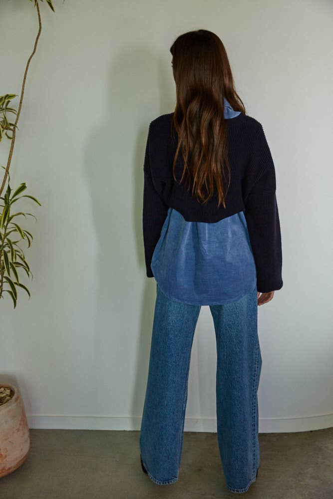 Person wearing a blue apron and dark jacket standing against a white wall with a plant to the left.