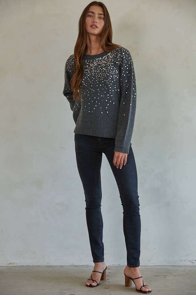 Woman wearing a gray sweater with sequin details and black pants against a plain background