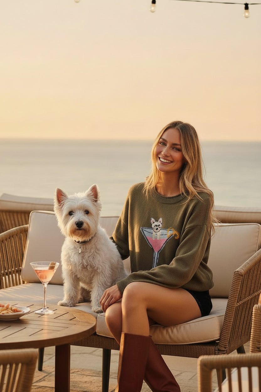 A woman modeling an olive green crewneck sweatshirt with a colorful cocktail graphic on the front, paired with dark denim shorts and brown leather knee-high boots, against a solid gray studio background.