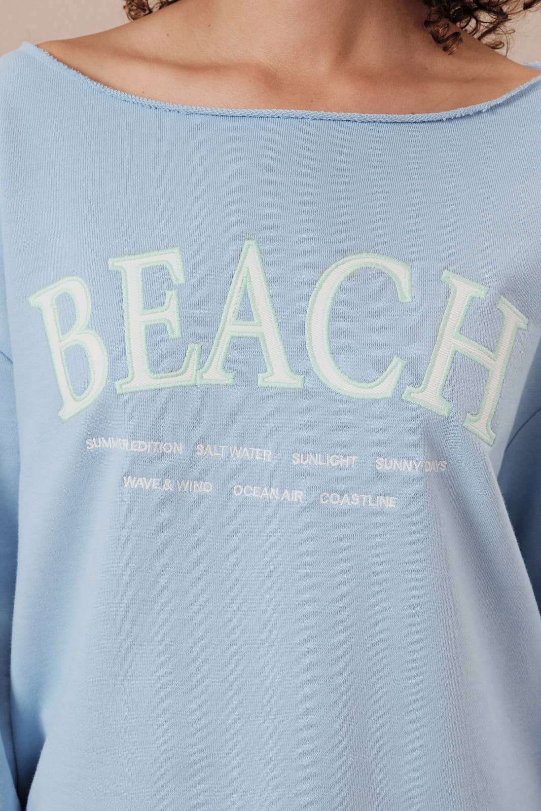 Light blue sweatshirt with 'BEACH' text and related words on a beige background