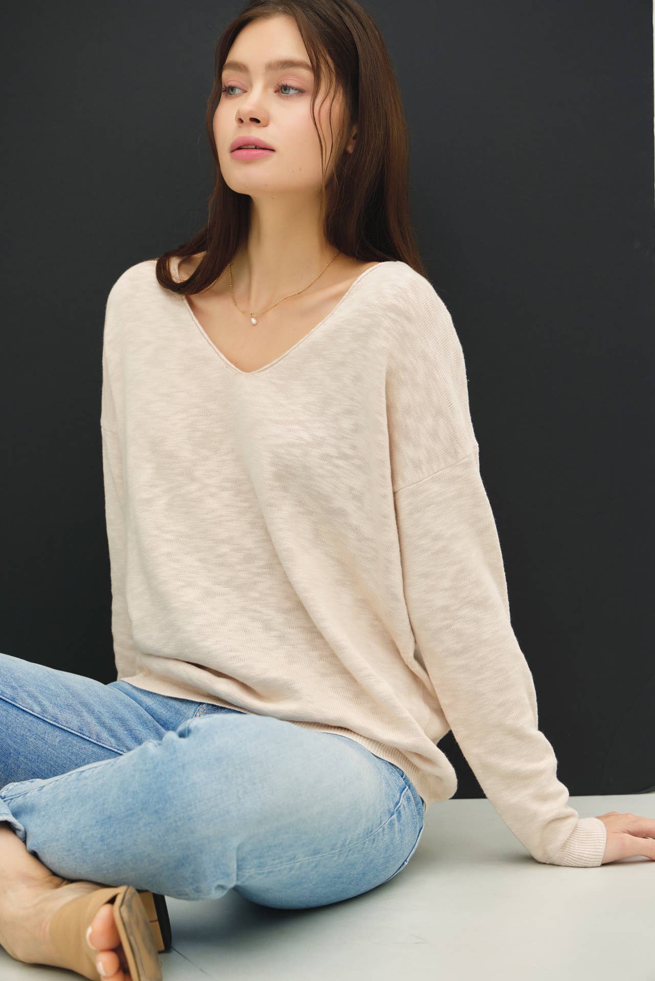 Woman wearing a beige sweater and blue jeans sitting on a gray surface with a black background