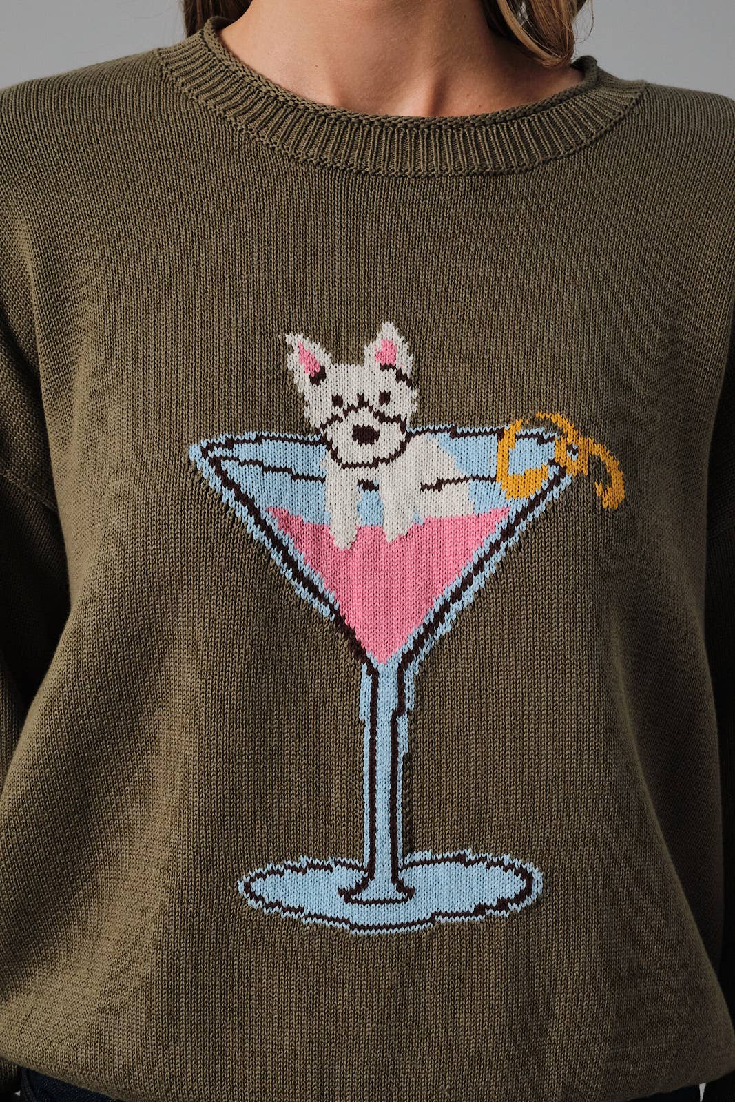 An olive green knit sweater featuring a knitted graphic of a white dog sitting in a pink martini glass with a yellow twist on the garnish.