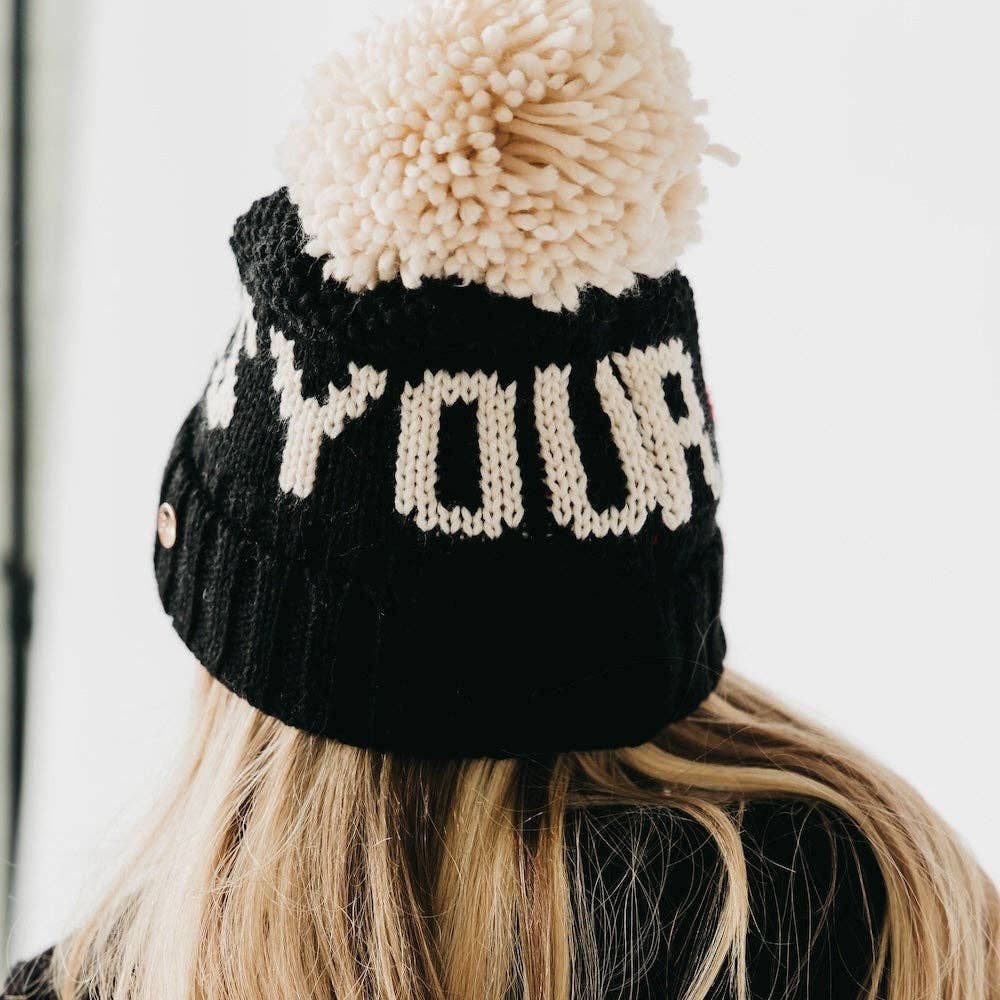 Black knit beanie with beige pom-pom and 'YOUR' text on a blurred background