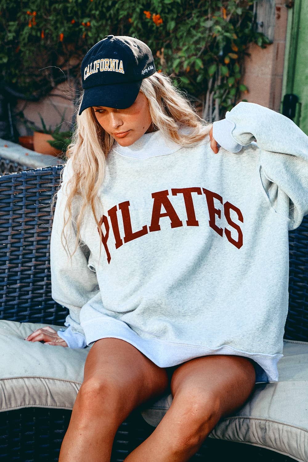 Person wearing a white 'PILATES' sweatshirt and black cap with 'California' text, sitting outdoors.