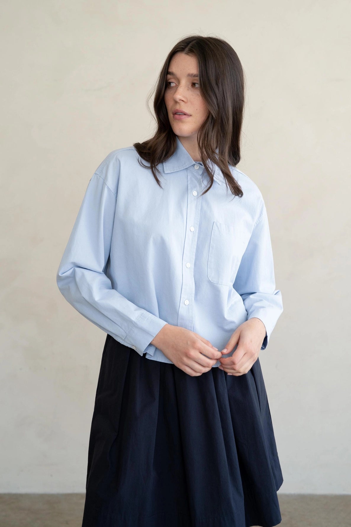 Woman wearing a light blue shirt and dark navy skirt against a plain background