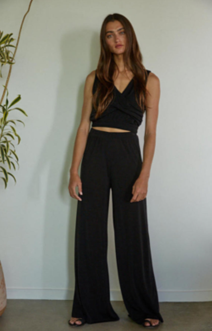 Woman wearing a black sleeveless top and wide-leg pants against a white wall.