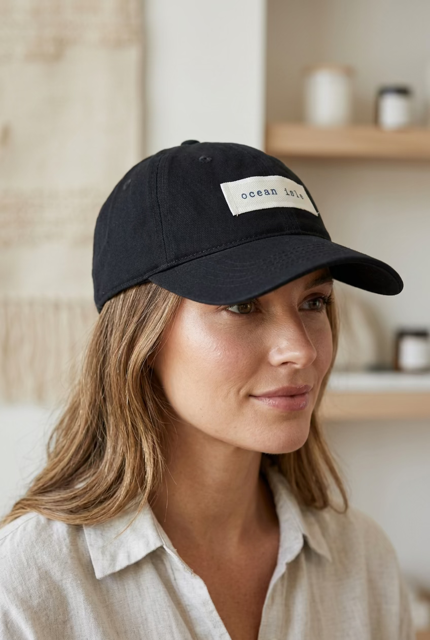 A black baseball cap with a beige fabric label reading 'ocean isle' in black text, photographed in a clothing store with racks of garments blurred in the background.