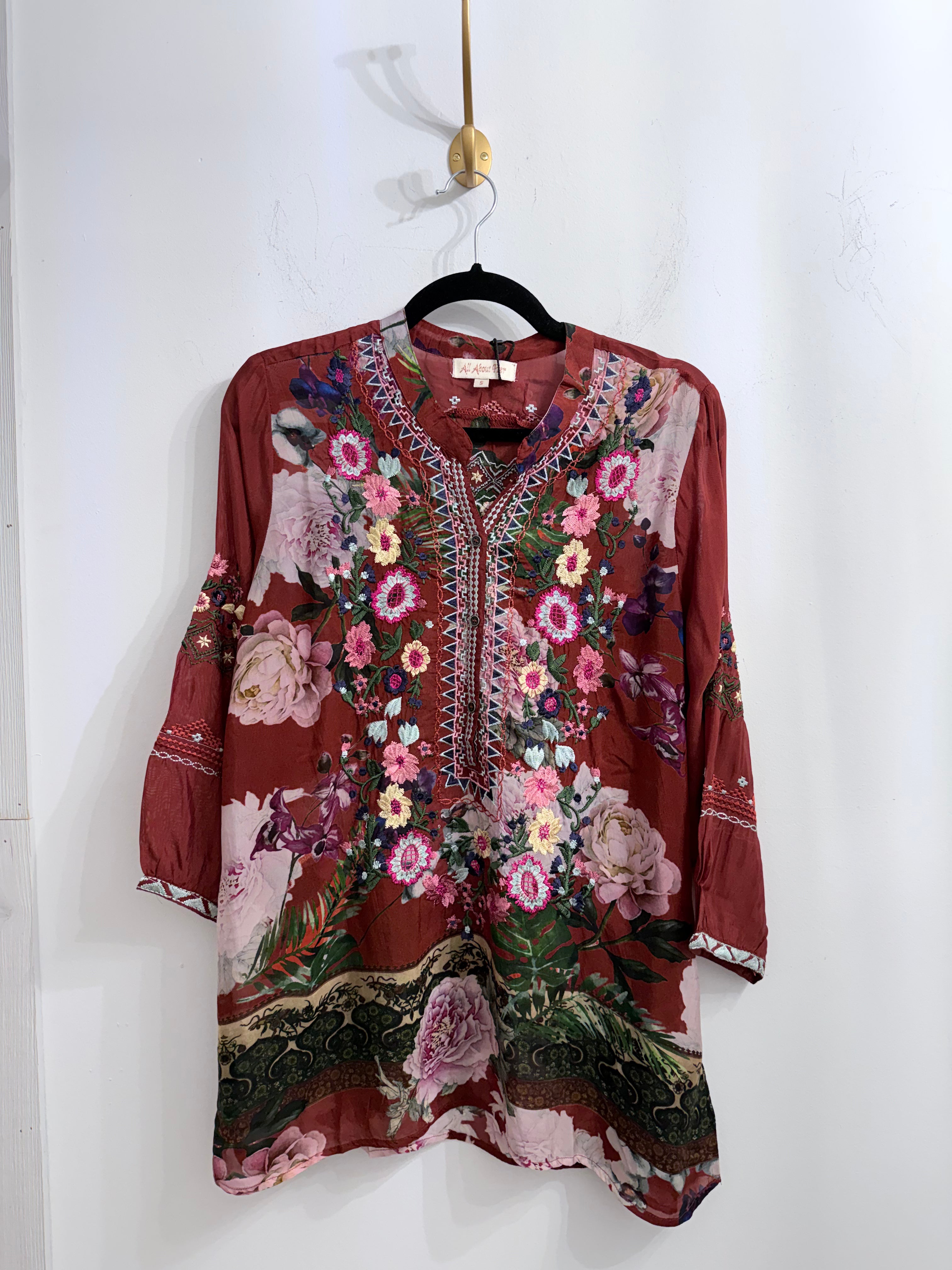 A long-sleeve tunic top with a reddish-brown base and a vibrant floral print, hanging on a black hanger against a white wall.