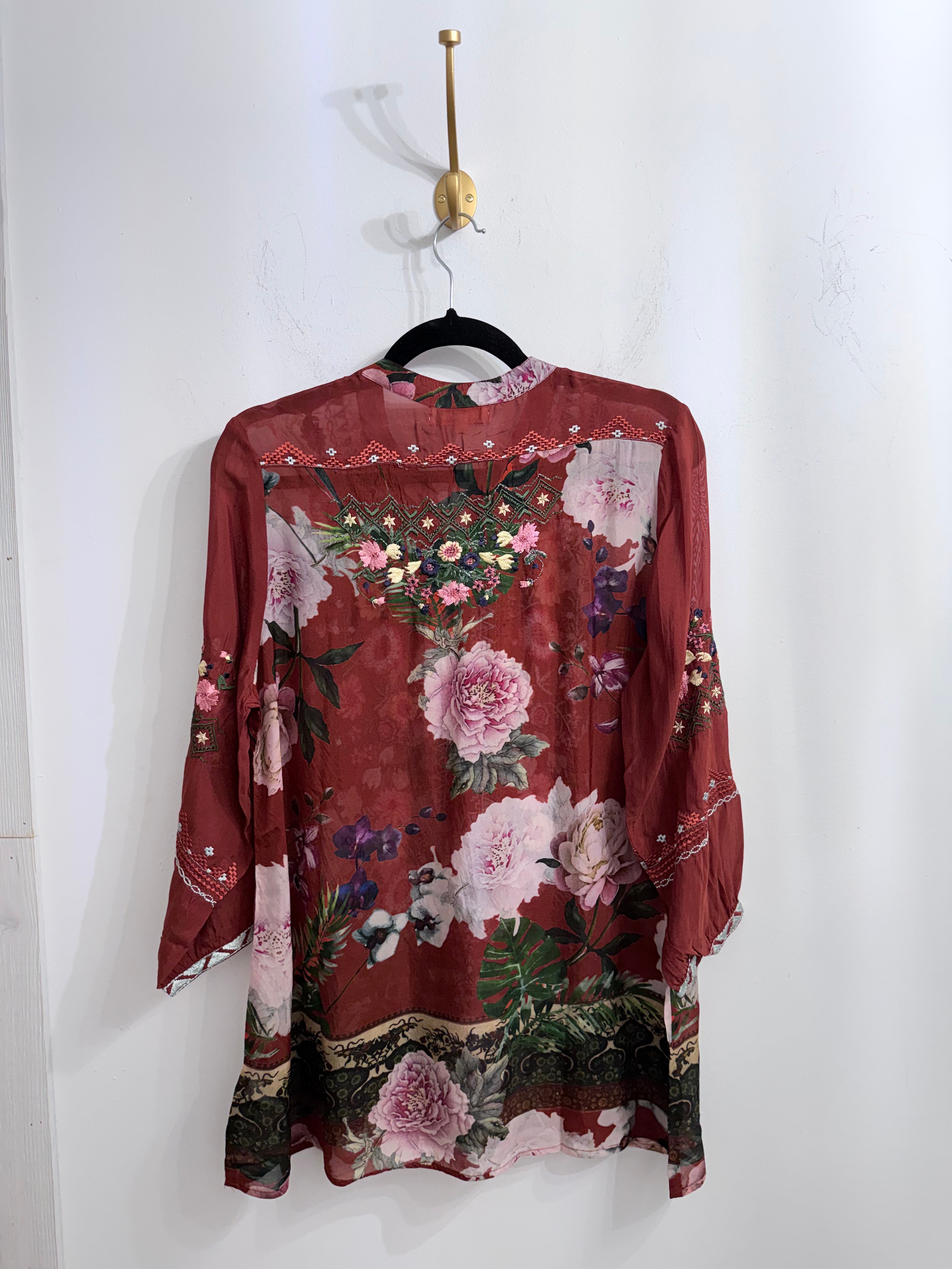 A long-sleeve tunic blouse with a red base and a large pink and green floral print, hanging on a black hanger against a white wall.