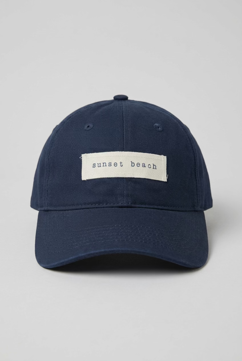 A dark navy blue baseball cap with a beige woven label reading 'sunset beach' in navy lettering, photographed inside a clothing store with racks of garments blurred in the background.