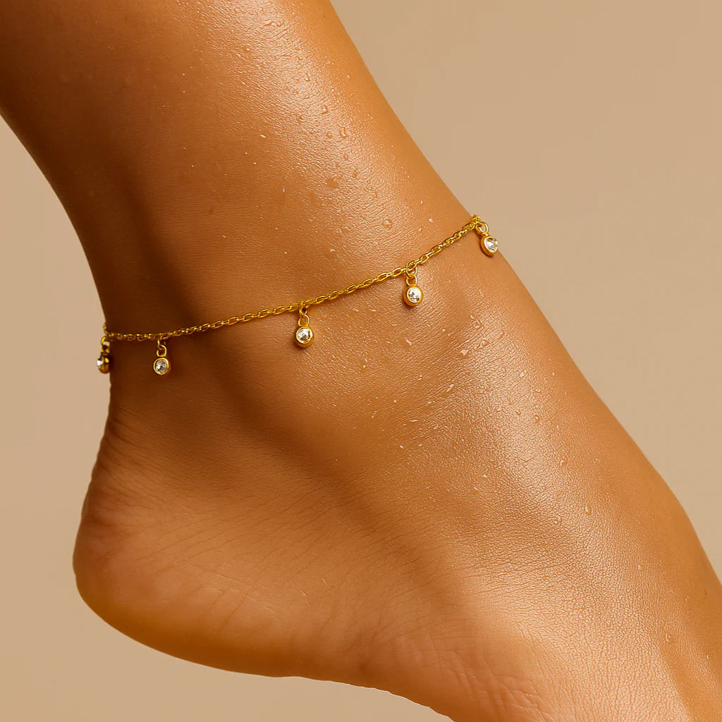 Gold anklet with small beads on a person's ankle against a beige background