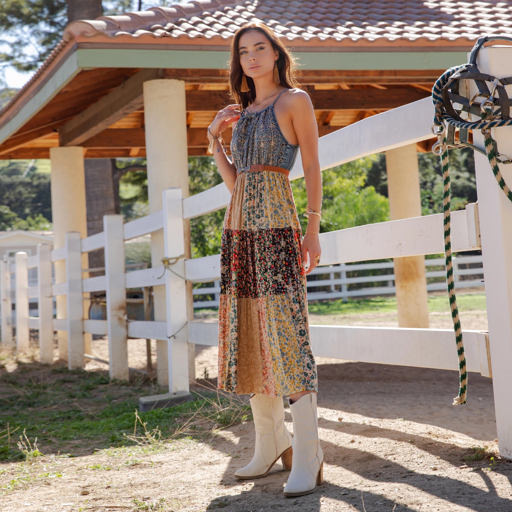 Woman in a patterned dress and white boots standing in front of a wooden building with a white fence.