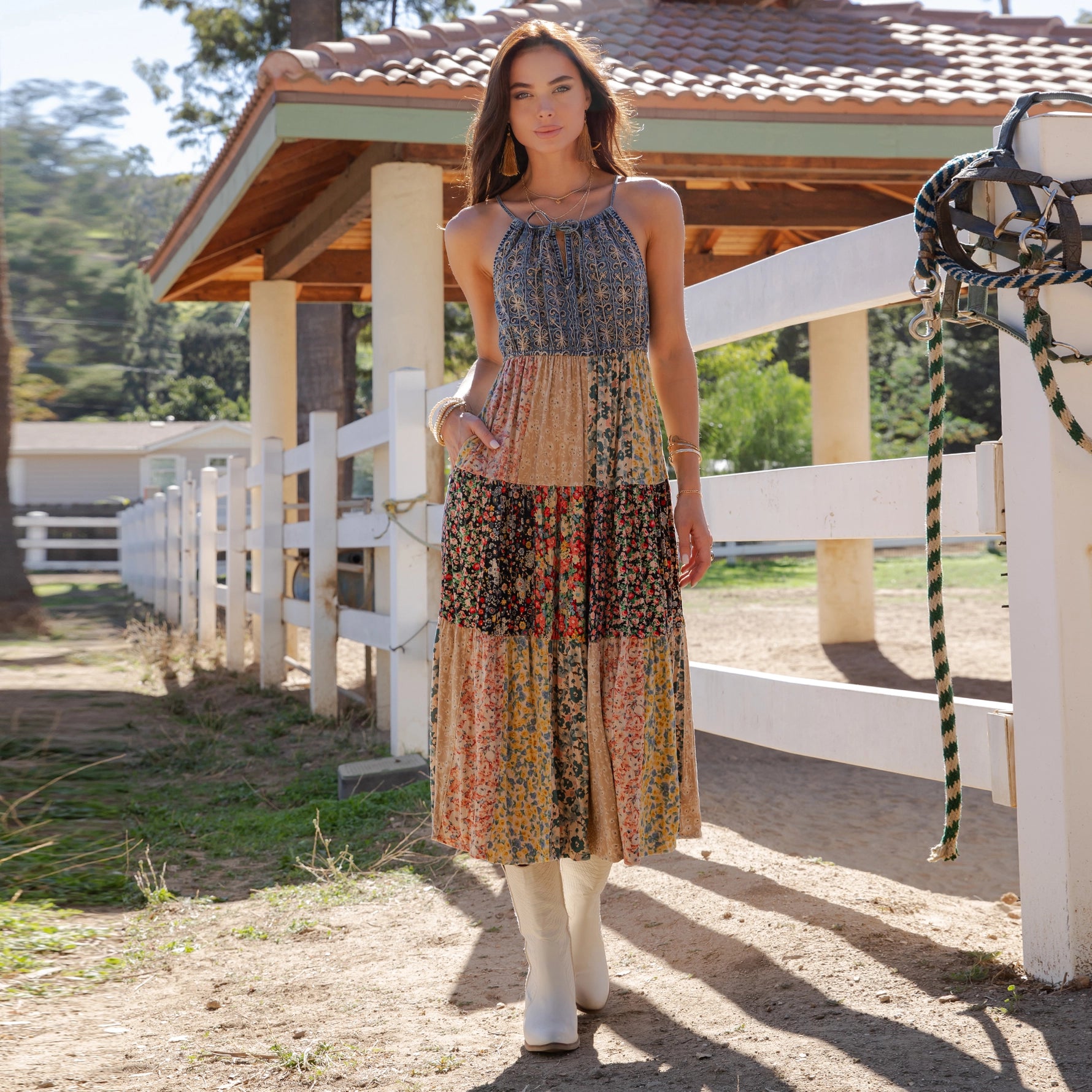 Woman in a patterned dress standing in front of a white fence with a wooden structure in the background.