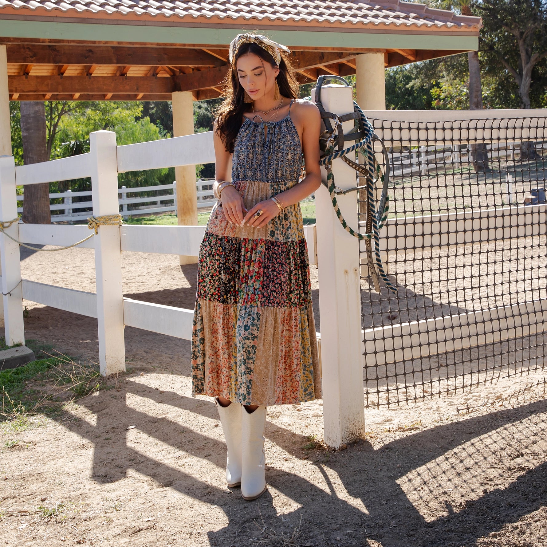 Woman in a patterned dress and white boots standing near a white fence with a wooden structure in the background.