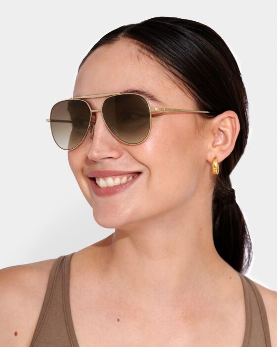 Woman wearing gold aviator sunglasses and a brown tank top against a white background