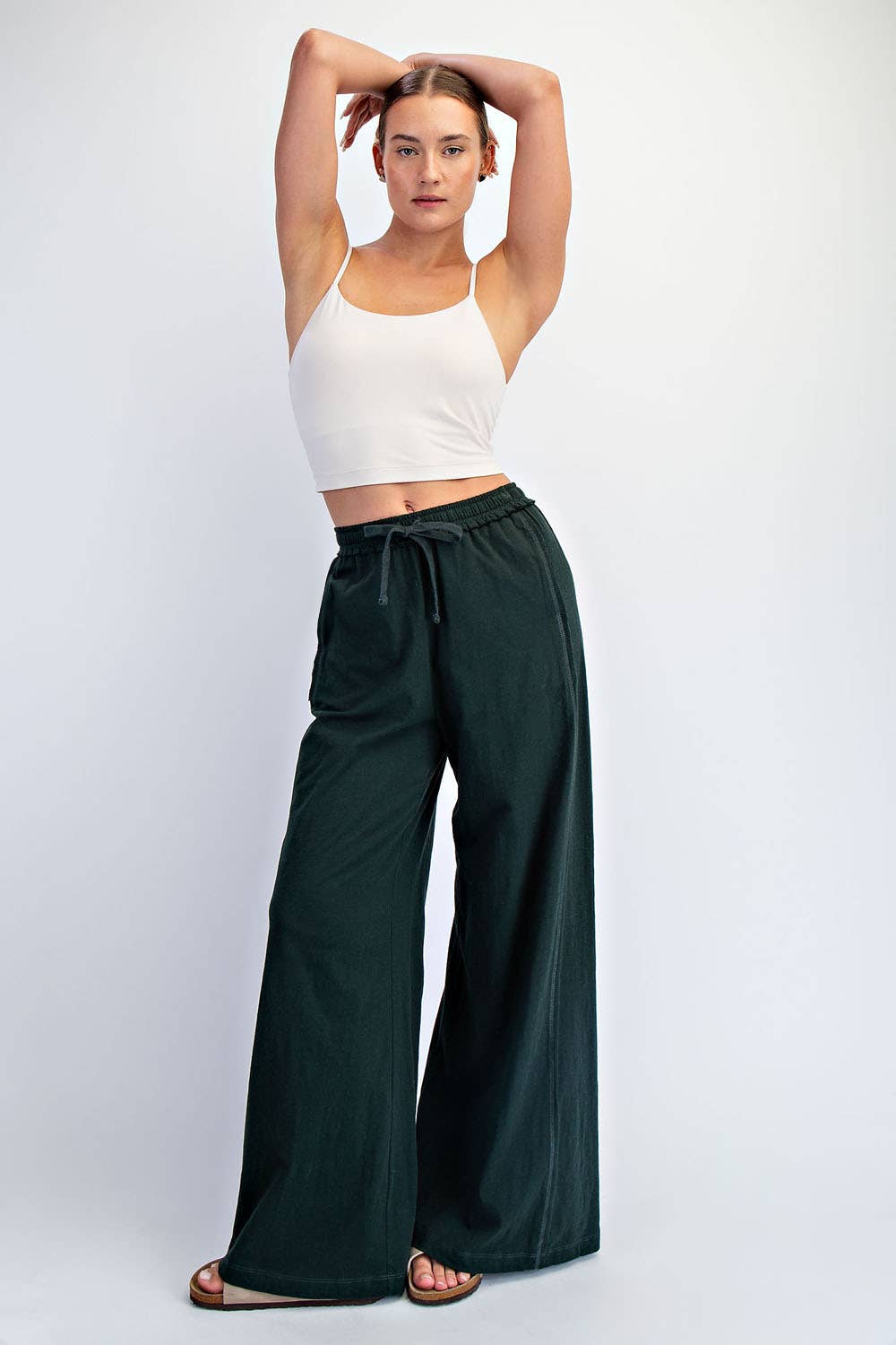 Woman wearing a white tank top and dark green wide-leg pants on a white background
