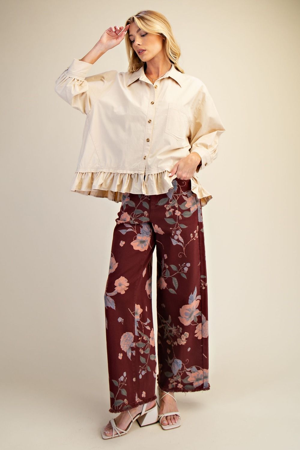 Woman wearing a cream blouse with ruffled details and burgundy floral pants on a beige background
