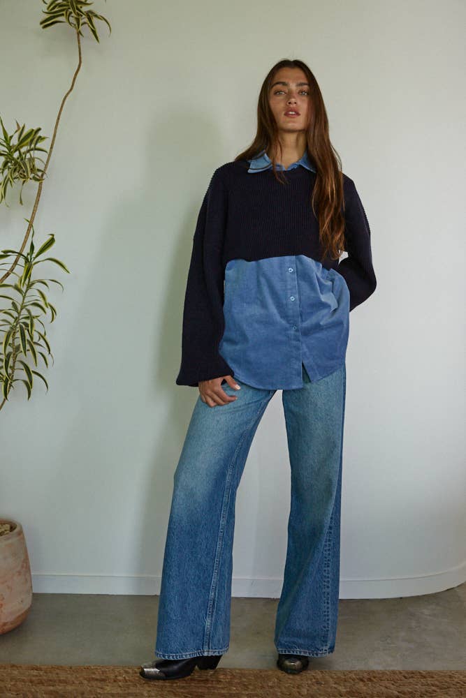 Person wearing a black sweater and blue denim outfit standing against a white wall with a plant on the left.